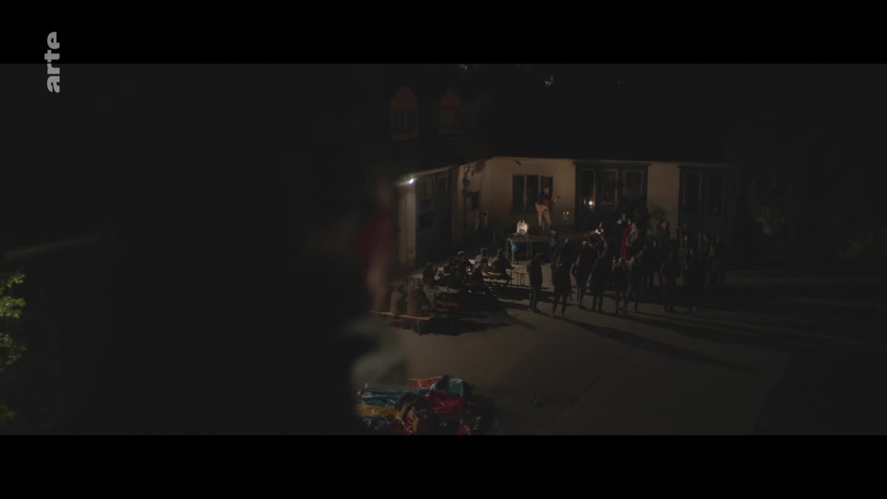 A line of riders on horseback slowly advances down the street, their silhouettes distinct against the lights from a house. People are gathered on the porch, watching.

