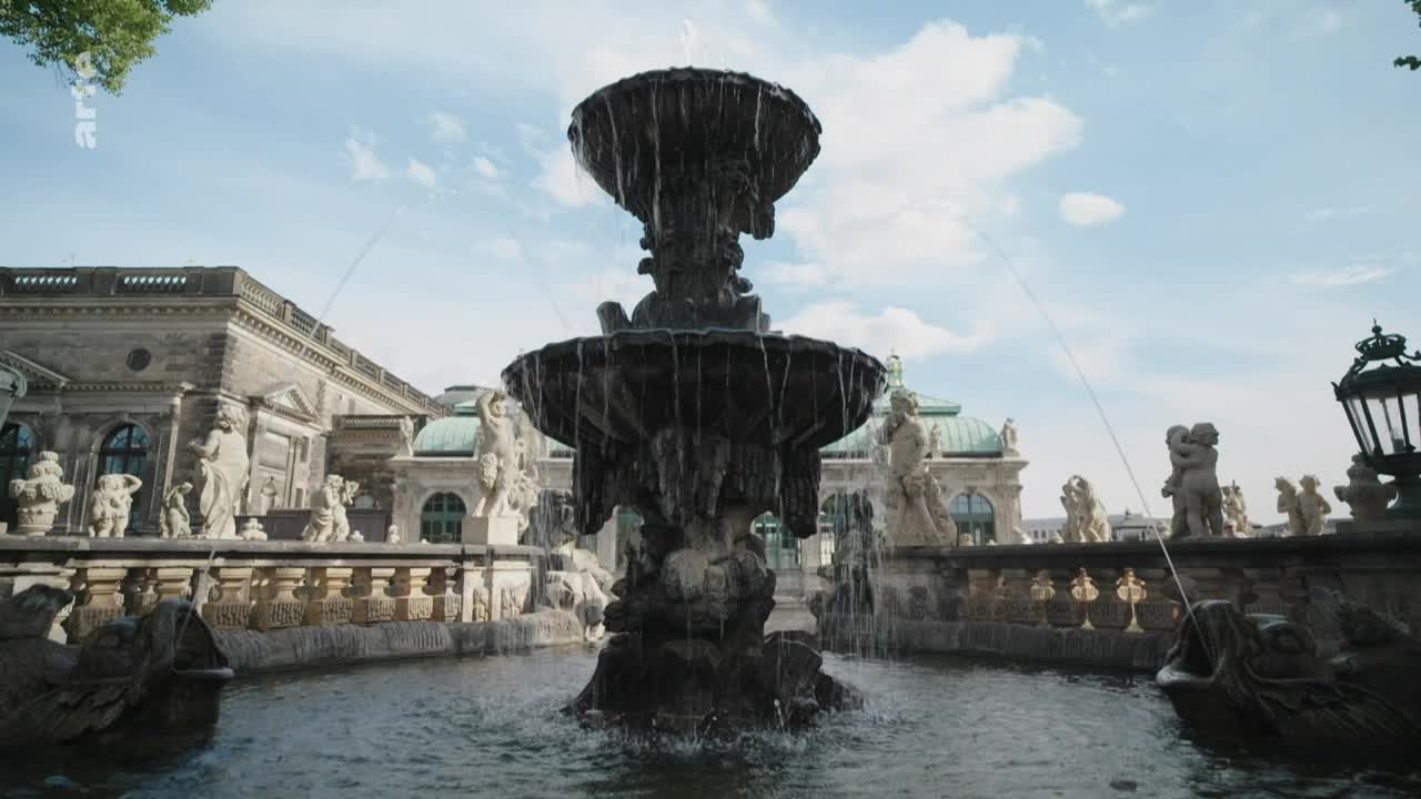 Water streams arch from the fountain's upper tiers, cascading into the pool below. Carved figures stand guard around the basin, while the sky above is a bright, summery blue.
