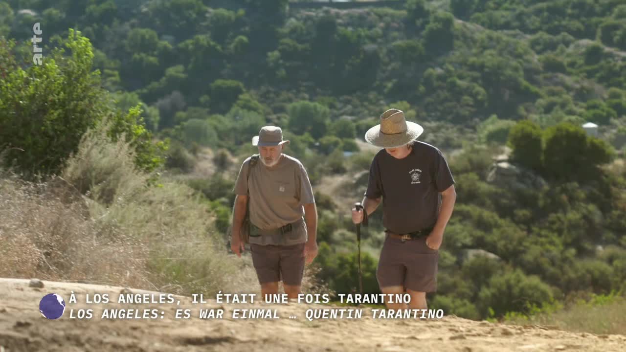 Two men in hats and shorts are hiking up a dirt path, the man on the right using a walking stick. The logo of a French-German TV channel, arte, is visible in the top left corner.
