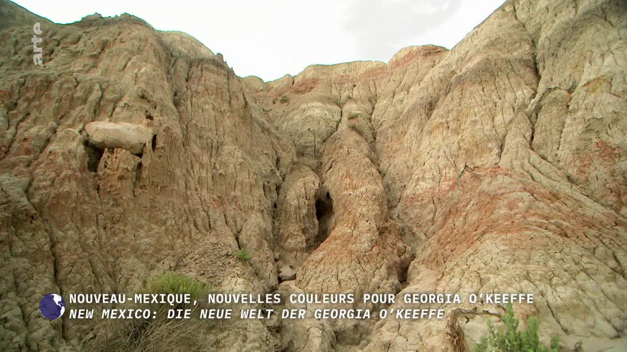 The eroded rock formations rise up, layered with streaks of reddish-brown and beige. A small cave-like opening is visible in the center of the canyon.
