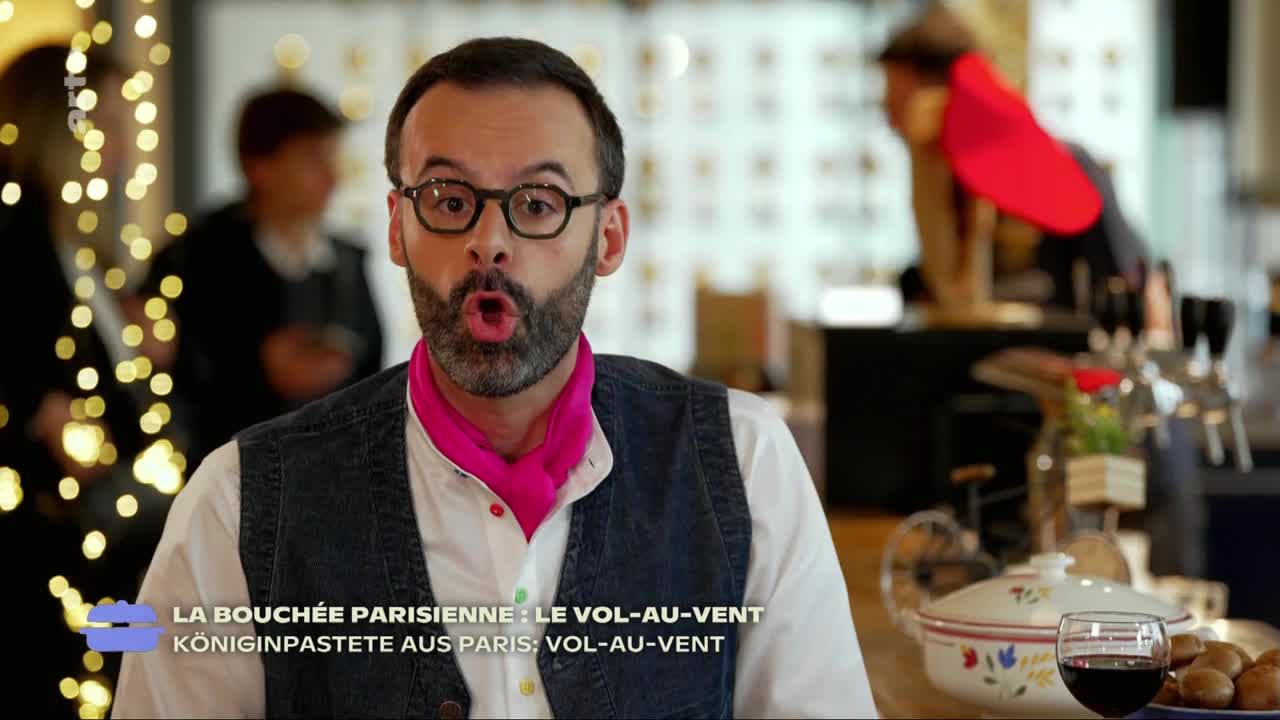 A man in a vest and glasses, with a pink scarf, is speaking directly to the camera, his mouth wide open. Behind him, a blurred background suggests a restaurant or café setting with a glimpse of food and drink.
