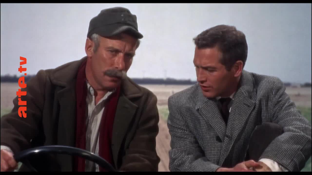 The older man, with a thick mustache, grips the steering wheel, while the younger man, in a checkered coat, leans in to listen. They're on a tractor, with a vast, flat landscape stretching out behind them.
