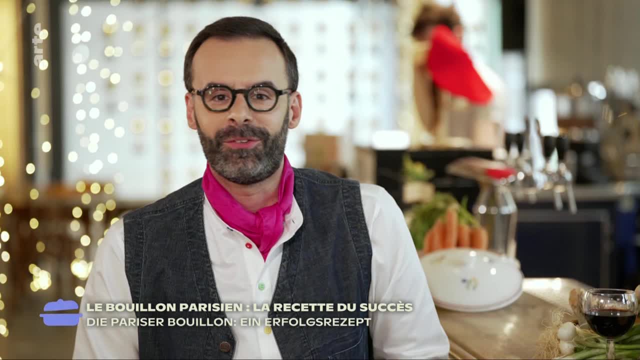A man with glasses and a pink scarf is speaking directly to the camera, the backdrop suggesting a kitchen or restaurant setting. Behind him, someone is holding a red cloth, possibly wiping something down.
