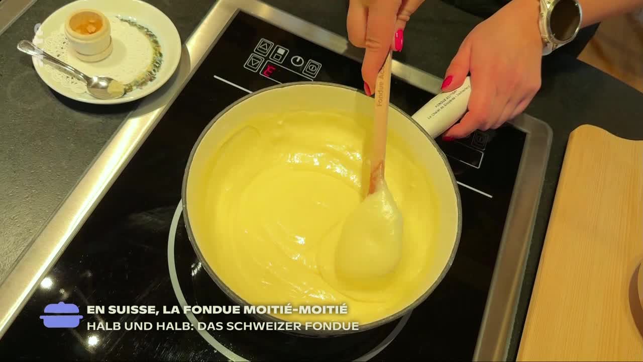 A wooden spoon swirls the creamy, yellow fondue in a pot on a stovetop.  The scene is from a cooking segment on arte, with a small plate of condiments nearby.
