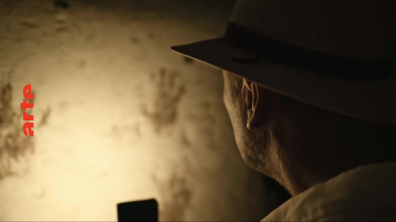 A man in a fedora peers at a wall marked with faint handprints. The red "arte" logo stands out against the muted tones.