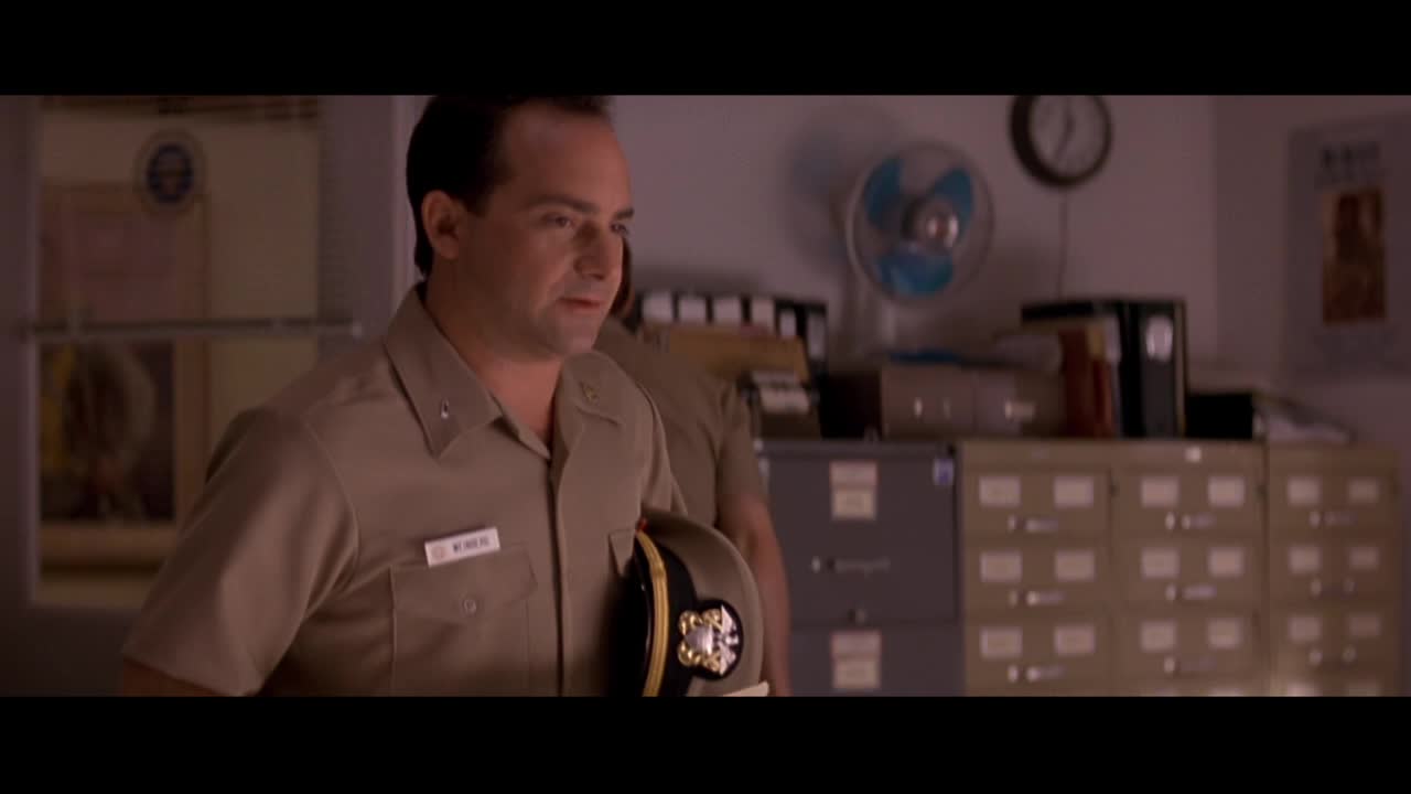 A man in a tan uniform holds a cap, its gold braid glinting. The room behind him is filled with filing cabinets and a whirring fan.