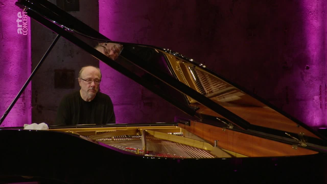 A man with glasses sits at a grand piano, his reflection visible in the lid. The stage is bathed in a soft, purple light.