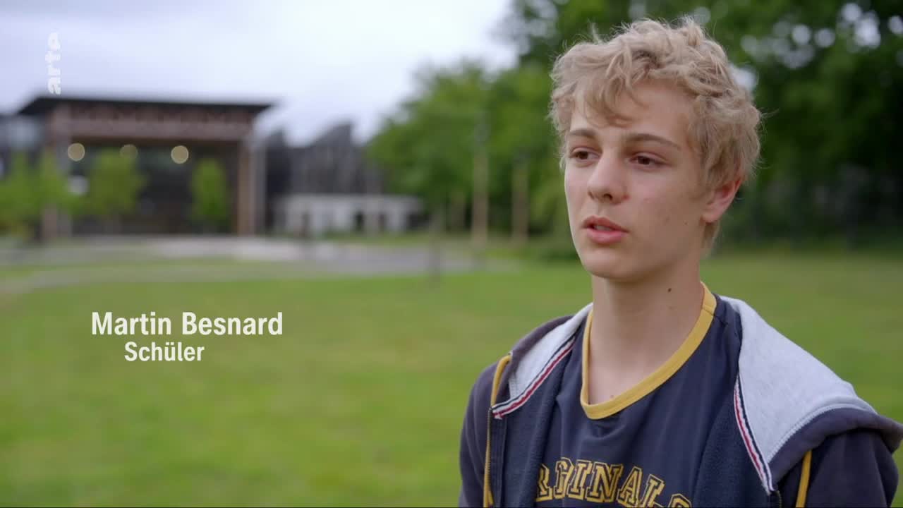 A young man with blond hair stands in a grassy area. Behind him, a building with a distinctive roof structure is visible. The arte logo appears in the upper left corner.
