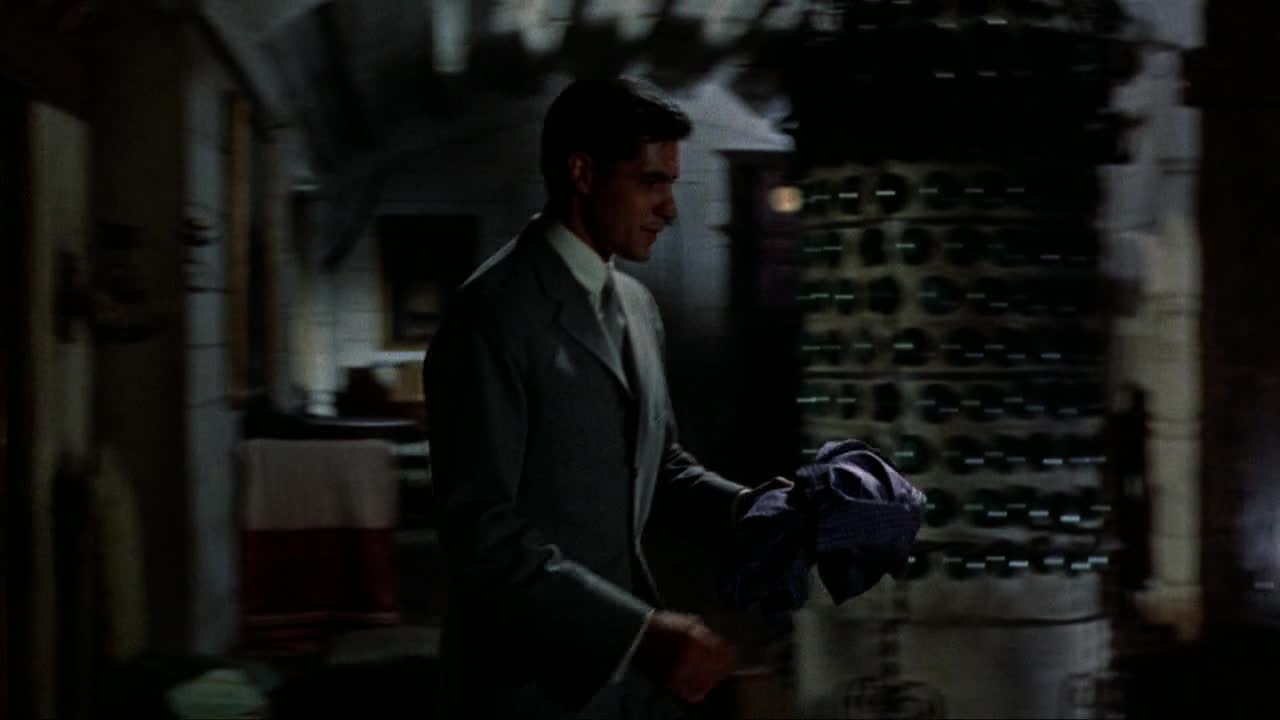 A man in a suit strides past a wall of wine bottles, his hand clutching a dark cloth. The dim light catches the sheen of his jacket as he moves quickly through the space.