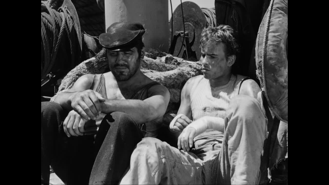 Two men, their faces smudged with dirt, sit slumped against a life preserver. One wears a cap, the other has a bandaged hand.