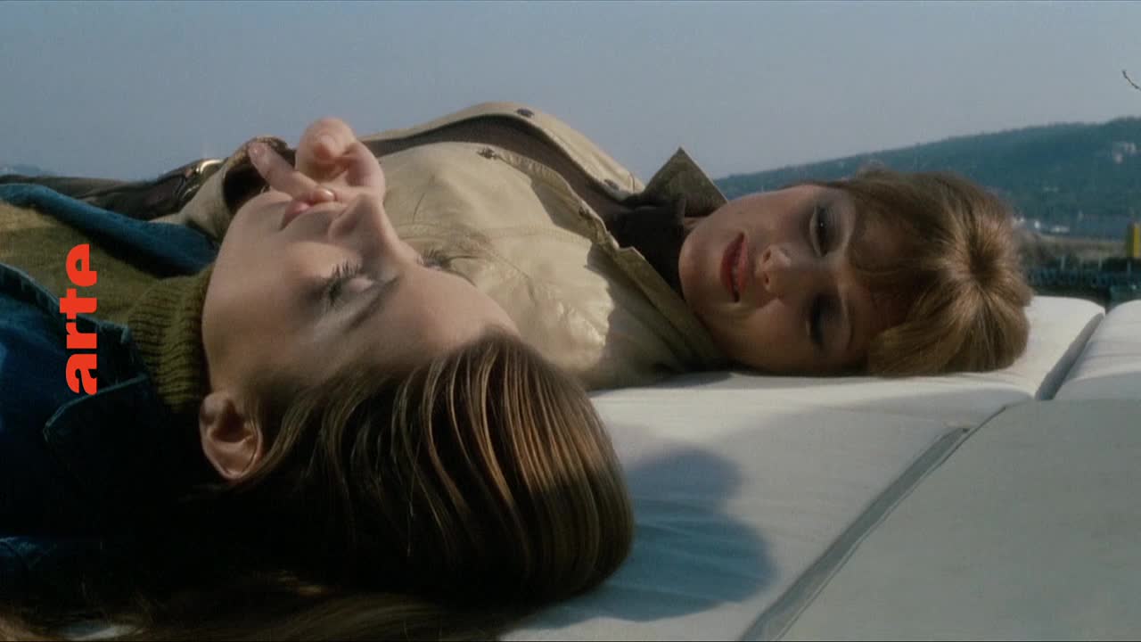 Two women lie on their backs, their faces turned towards the sky. The one in the foreground rests her head on her arm, eyes closed, while the other looks up with a faint smile.