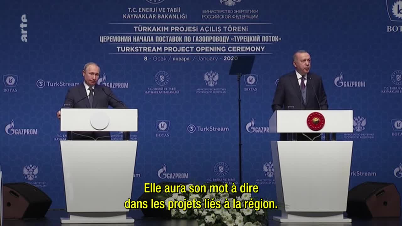 Two men stand at podiums, one speaking, the other listening intently. The backdrop is a deep blue, emblazoned with logos and text in multiple languages, hinting at an international energy announcement.