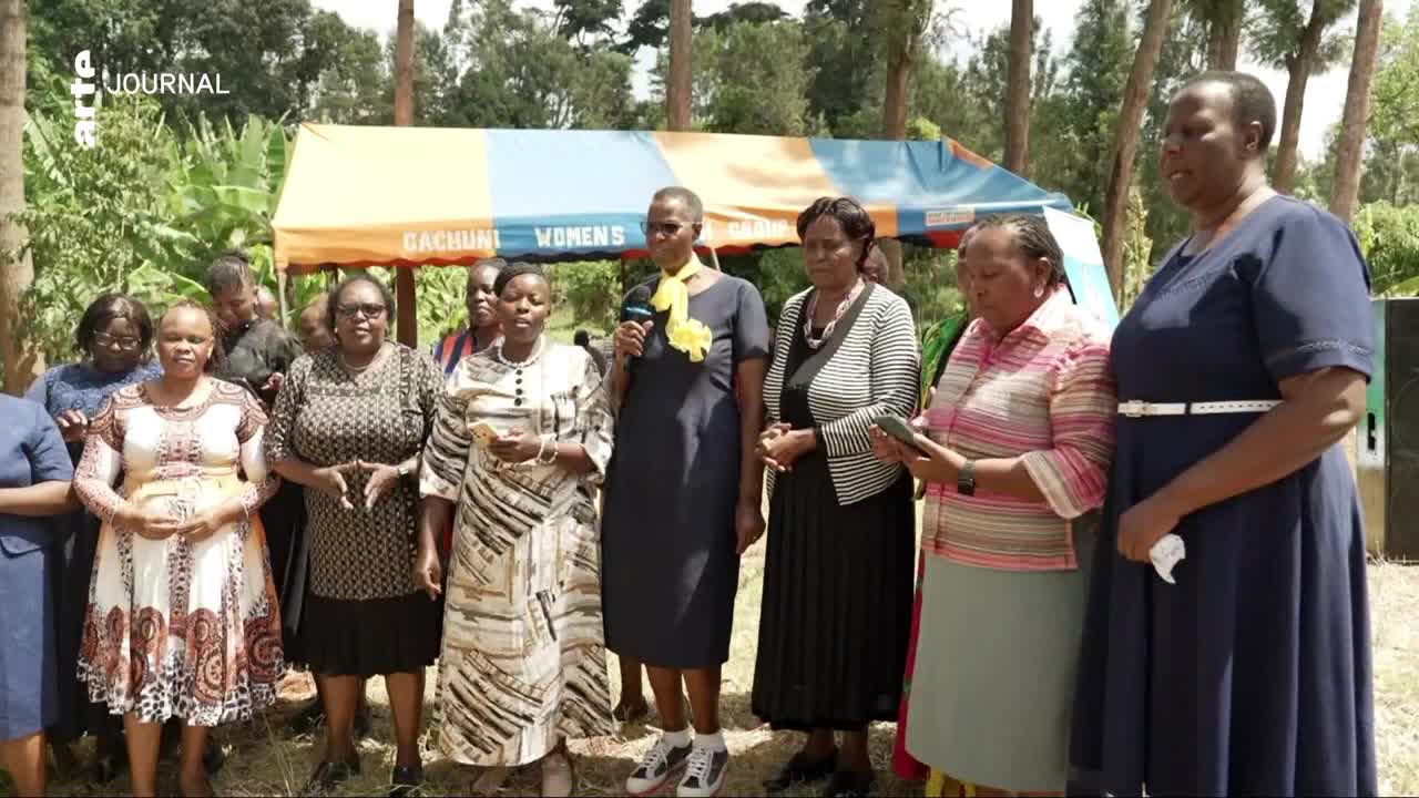 A group of women stands before a tent that reads "Gachuni Women's." One woman in the center holds a microphone, her yellow scarf bright against her dark dress. Another woman to her right consults a tablet.