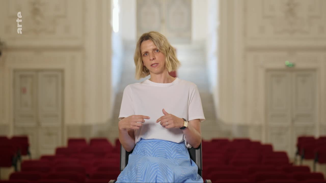 A woman with short blonde hair sits in a chair, gesturing with her hands. She wears a white t-shirt and a blue patterned skirt, her gaze directed forward. Behind her, rows of empty red seats stretch into the distance within a grand hall.