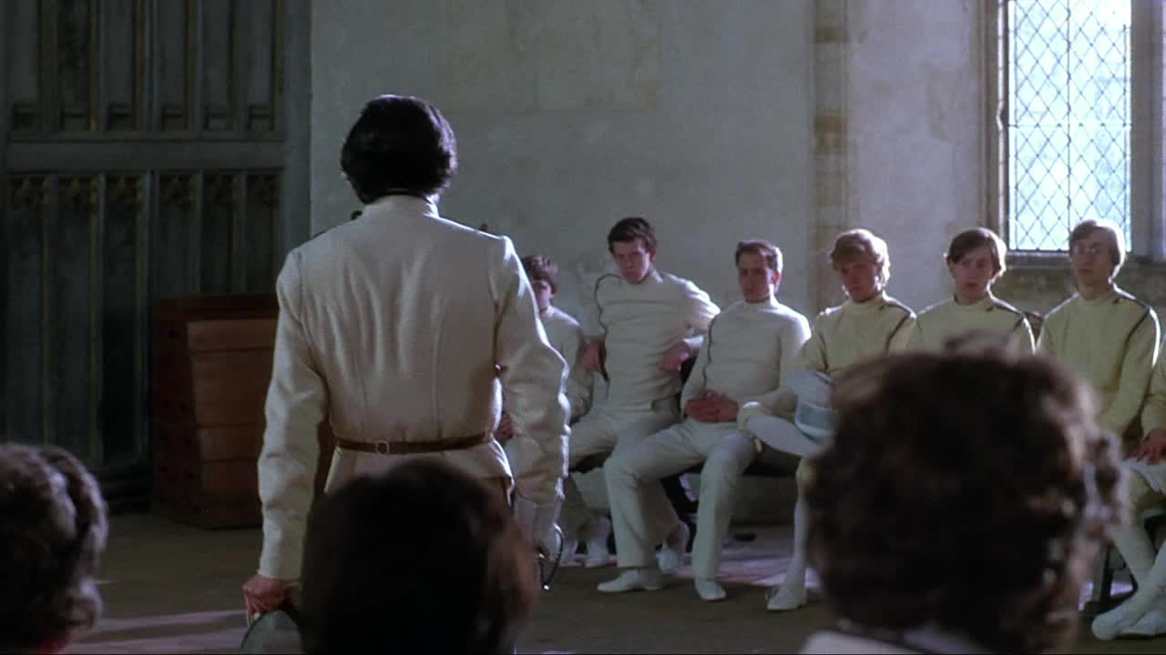 A man in a white fencing uniform stands facing a group of young men seated on a bench. The room, with its high windows and stone walls, feels like an old French academy.