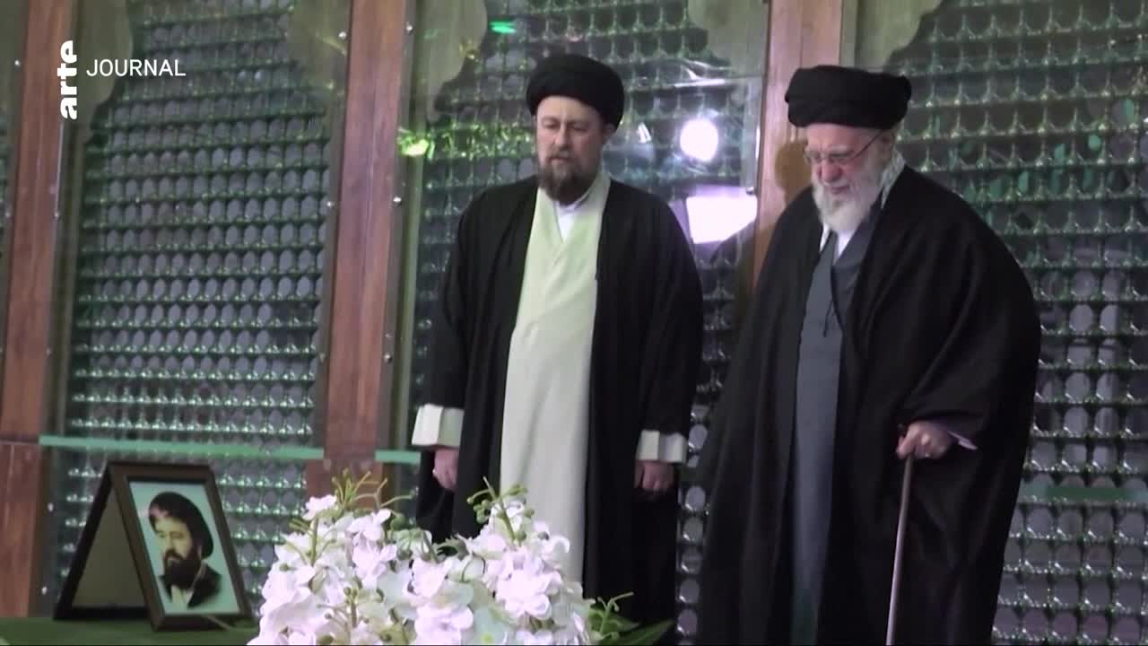 Two men in clerical robes stand before a framed photograph and a floral arrangement. The man on the right leans on a cane as he looks down.