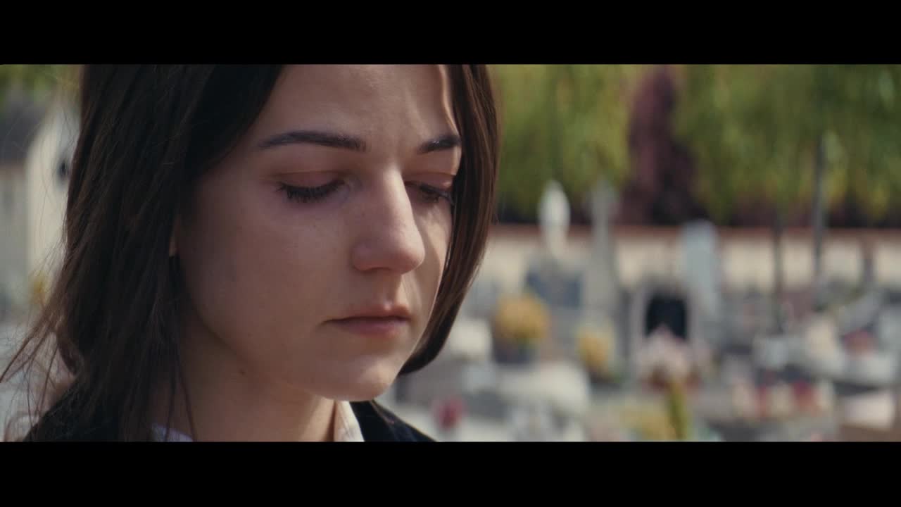 A young woman stands with her head bowed, her gaze fixed on the ground. Behind her, the blurred shapes of gravestones suggest a cemetery, a quiet place for reflection.