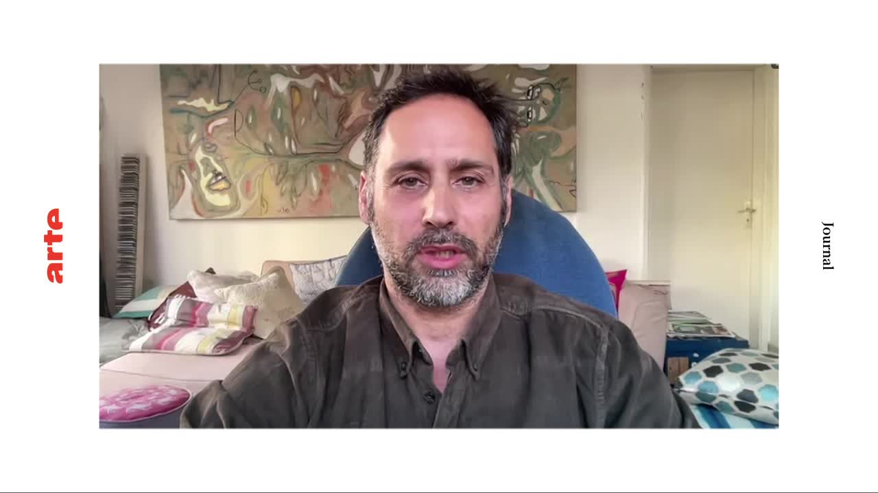 A man with a beard speaks directly to the camera from a comfortable setting. Behind him, a colorful, abstract painting hangs on the wall.