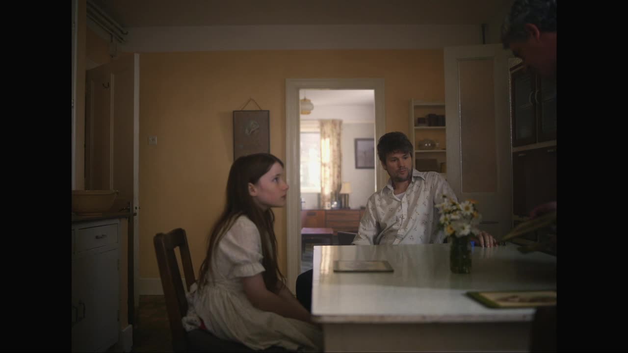 A young girl in a white dress sits at a table, her gaze fixed on something beyond the frame. A man in a patterned shirt leans forward, his hand resting near a small vase of daisies.