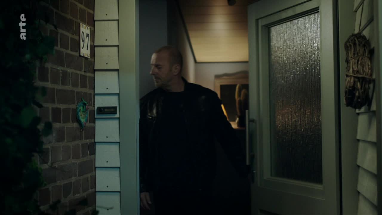 A man in a dark jacket steps through an open doorway, his gaze turned to the side. The brickwork beside him bears a house number and a curious green door knocker.