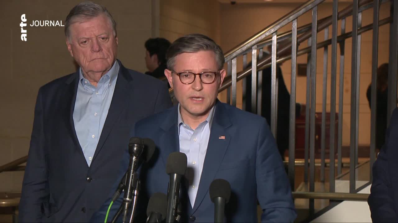 A man in a blue suit and glasses speaks into a microphone, while another man in a suit stands behind him. The arte journal logo appears in the upper left corner.