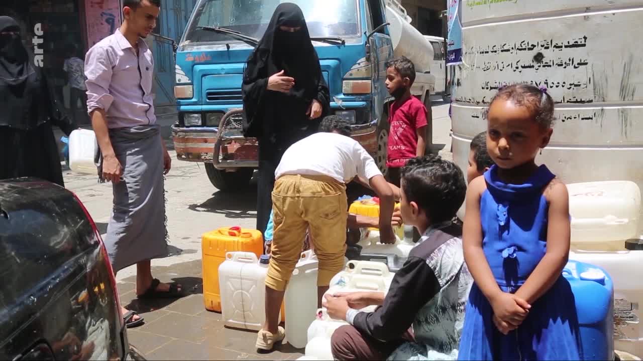 Children are filling large plastic containers with water from a truck. A young girl in a blue dress stands near a large white tank.