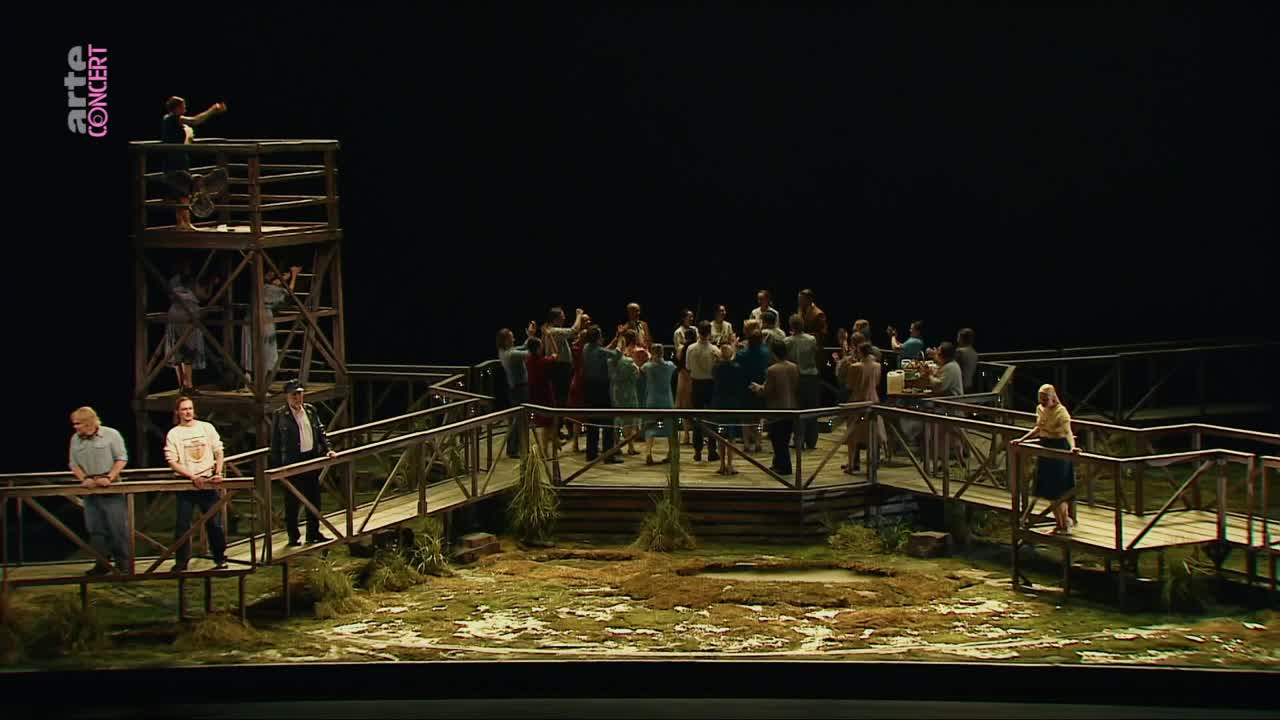A group of people gathers on a wooden platform, some leaning over railings. On a higher structure, a figure stands pointing outwards.
