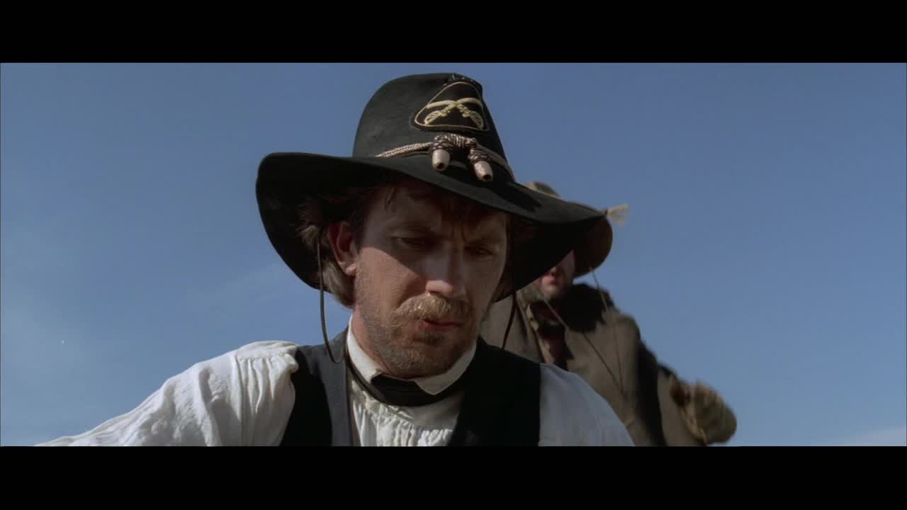 A man in a wide-brimmed hat, his face etched with concern, looks down as another figure, cloaked and indistinct, rides past. The vast, clear sky stretches above them.