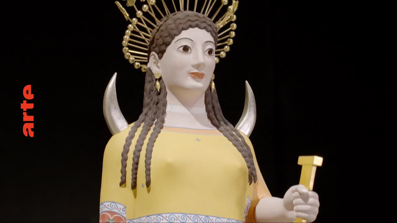 A statue of a woman, adorned with a golden headdress and crescent moons, stands before a black backdrop. Her yellow tunic is decorated with a band of blue and red patterns.