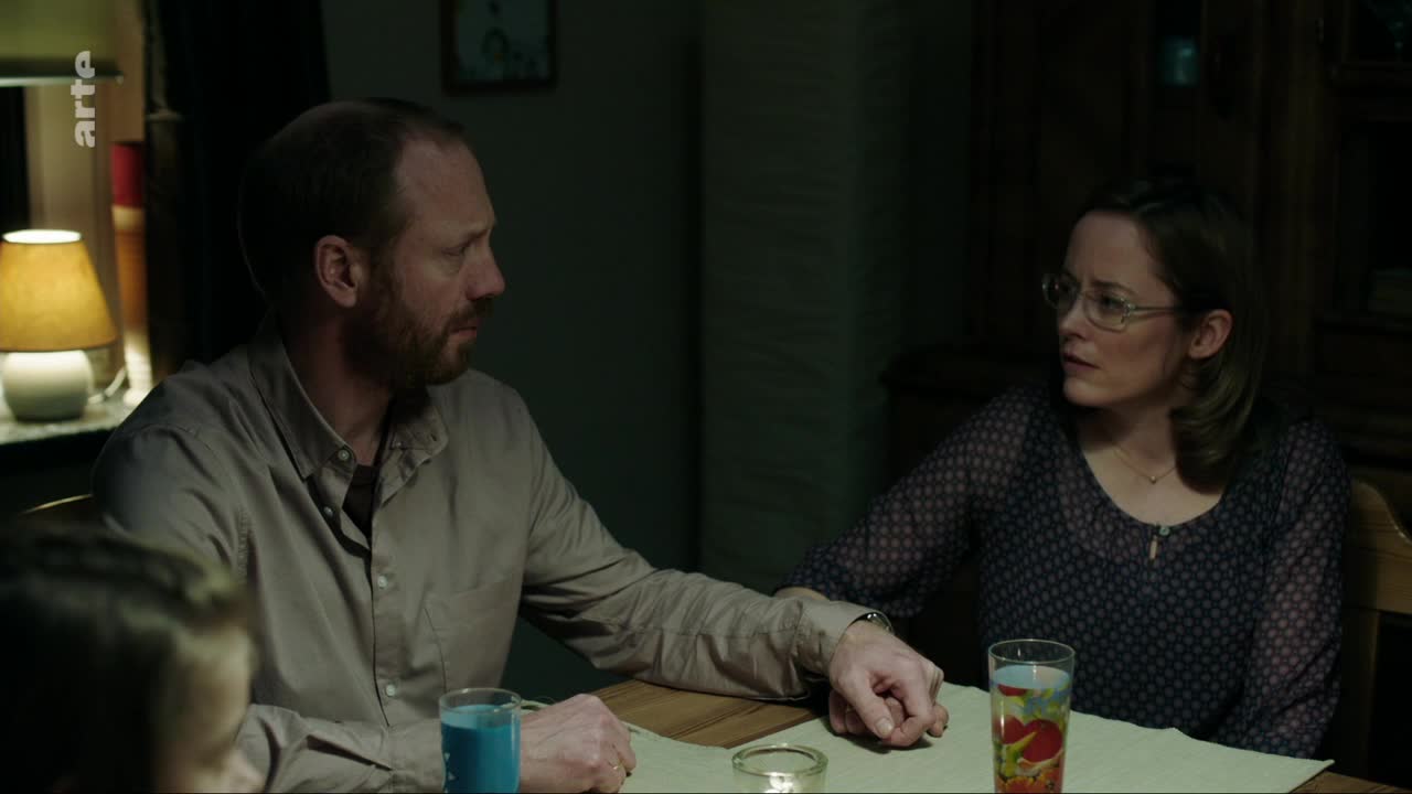 A man and a woman sit at a table, their faces illuminated by a warm lamp. The woman, wearing glasses, looks intently at the man, who is speaking with his hand resting on the table.