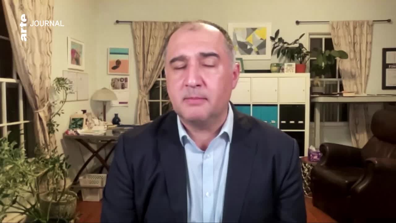 A man in a dark suit and light blue shirt is speaking, his eyes closed. Behind him, a wall displays abstract art, and a tall plant stands near a window.