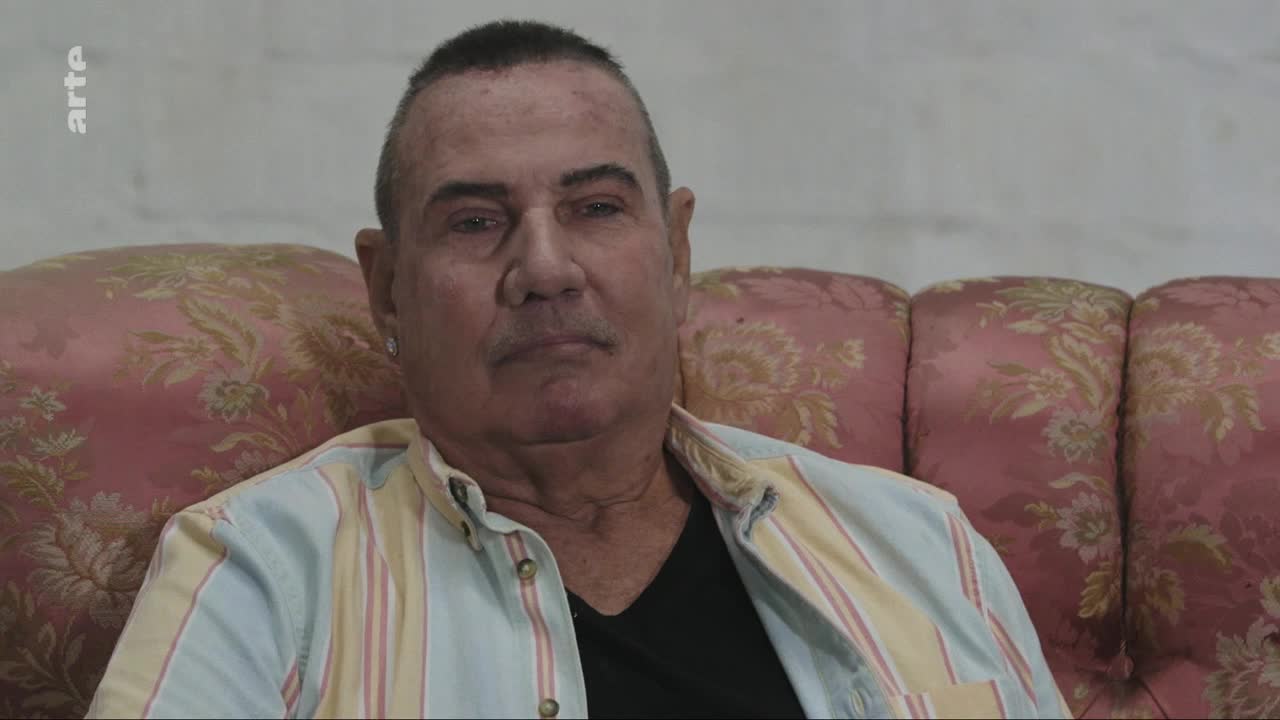 A man with short, dark hair sits on a pink floral sofa. He wears a striped shirt over a black t-shirt. The arte logo appears in the upper left corner.