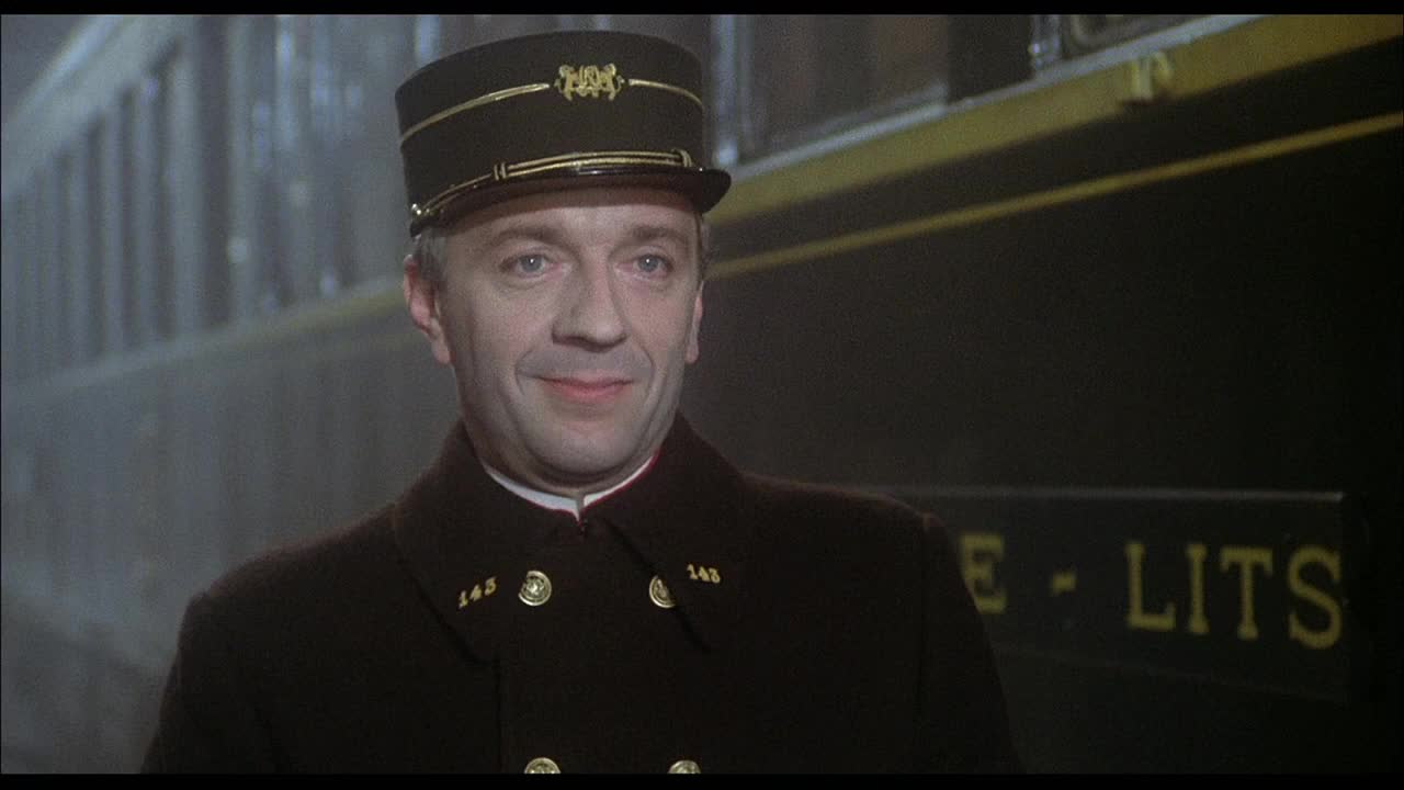 A train conductor stands by the dark carriages, a faint smile on his face. The lettering on the train car reads "VOYAGE - LITS," indicating a sleeping car.