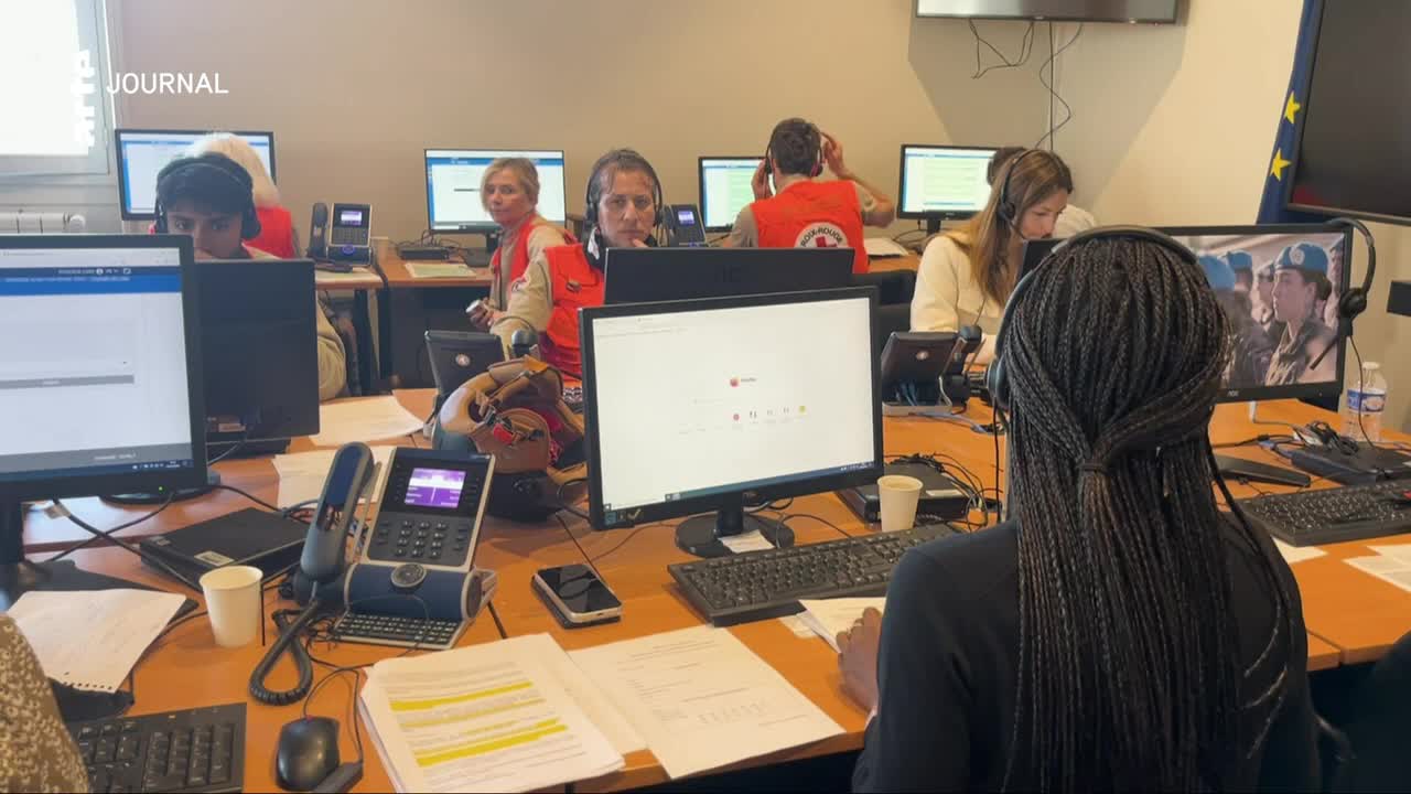 People in red vests are working at desks in a busy office. A woman in the foreground with braided hair looks at a screen showing a group of people in uniform.