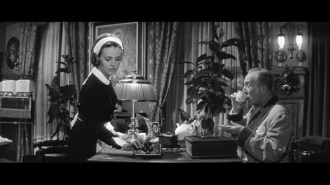 A young woman in a maid's uniform carefully arranges items on a desk. Across from her, a man in a robe sips from a teacup.