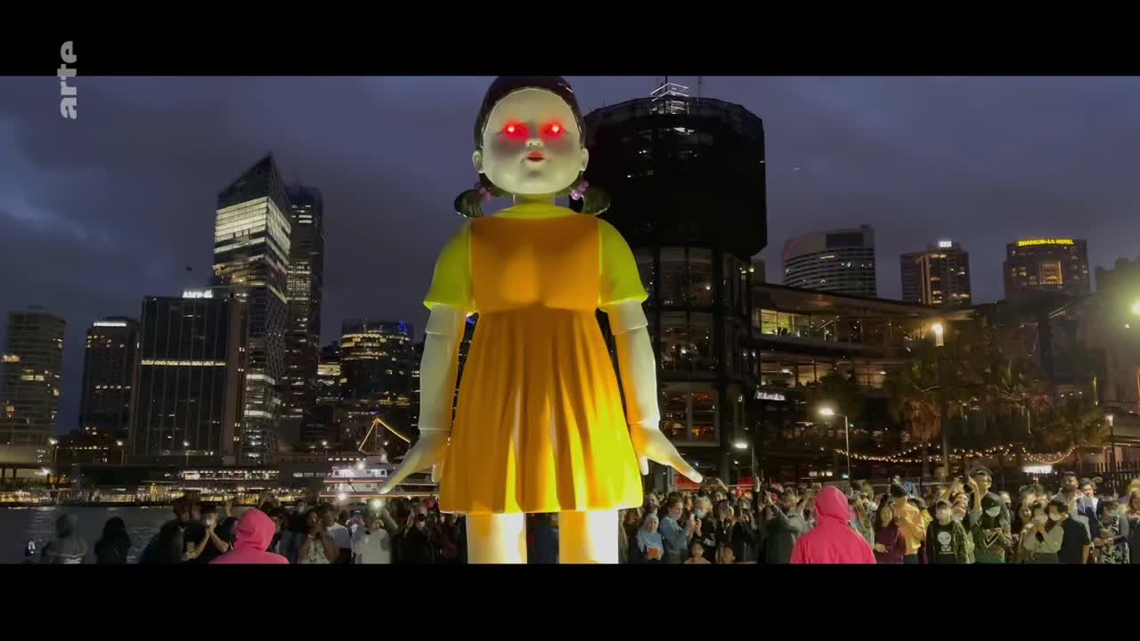 A giant doll with glowing red eyes stands in front of a city skyline at dusk. A crowd gathers, some in bright pink hoodies, to watch the spectacle.