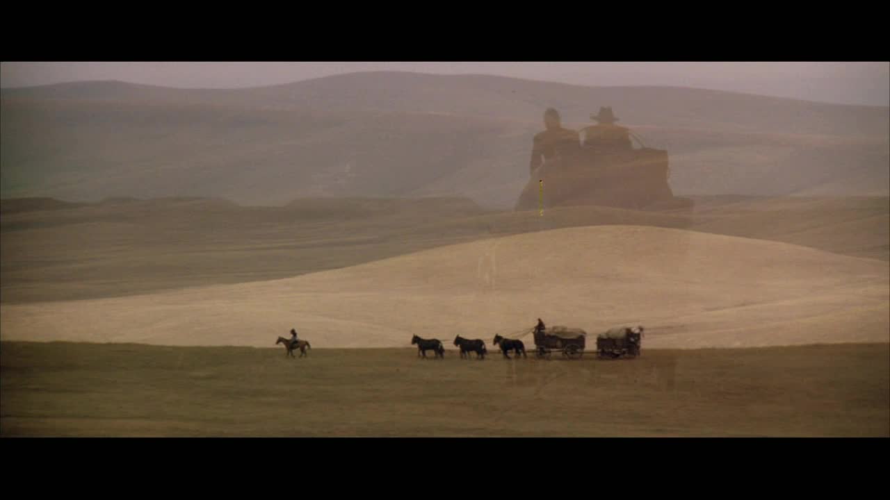 A lone rider on horseback moves across the vast, dusty plains. Behind them, a small wagon train, pulled by horses, makes its slow progress.