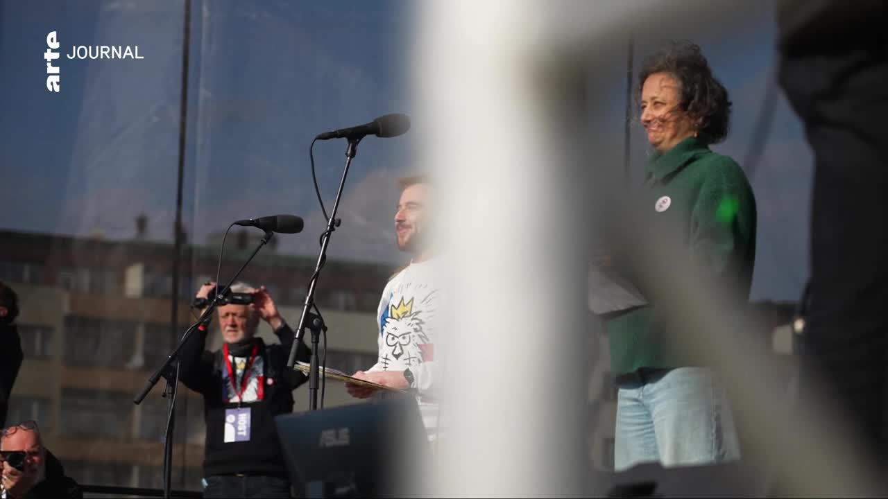 A woman in a green hoodie smiles, holding papers. A man in a white shirt with a graphic print stands nearby, holding a baton.