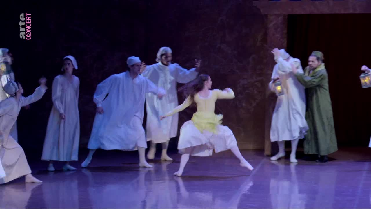 A young woman in a bright yellow tunic and white skirt leaps across the stage. Several figures in long white nightgowns, some holding lanterns, move around her.