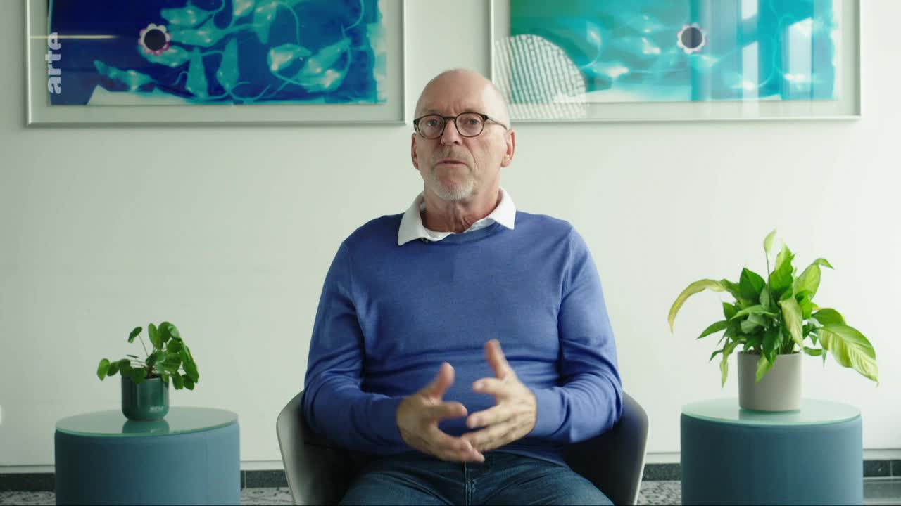 A man with glasses, wearing a blue sweater, speaks with his hands animated. Behind him, two framed artworks hang on the wall, one featuring the arte logo.