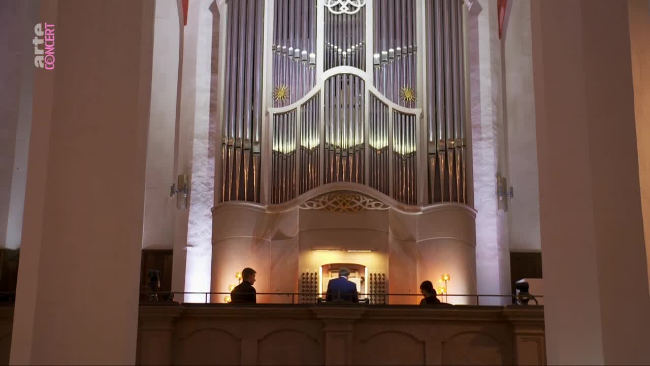 A man in a blue suit plays the massive organ, his hands moving over the keyboards. Two other figures stand beside him on the balcony, silhouetted against the warm glow of the pipes.
