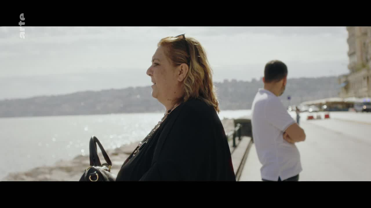 A woman with blonde hair, wearing a black shawl, stands by the water. A man in a white polo shirt stands further down the street, his arms crossed.