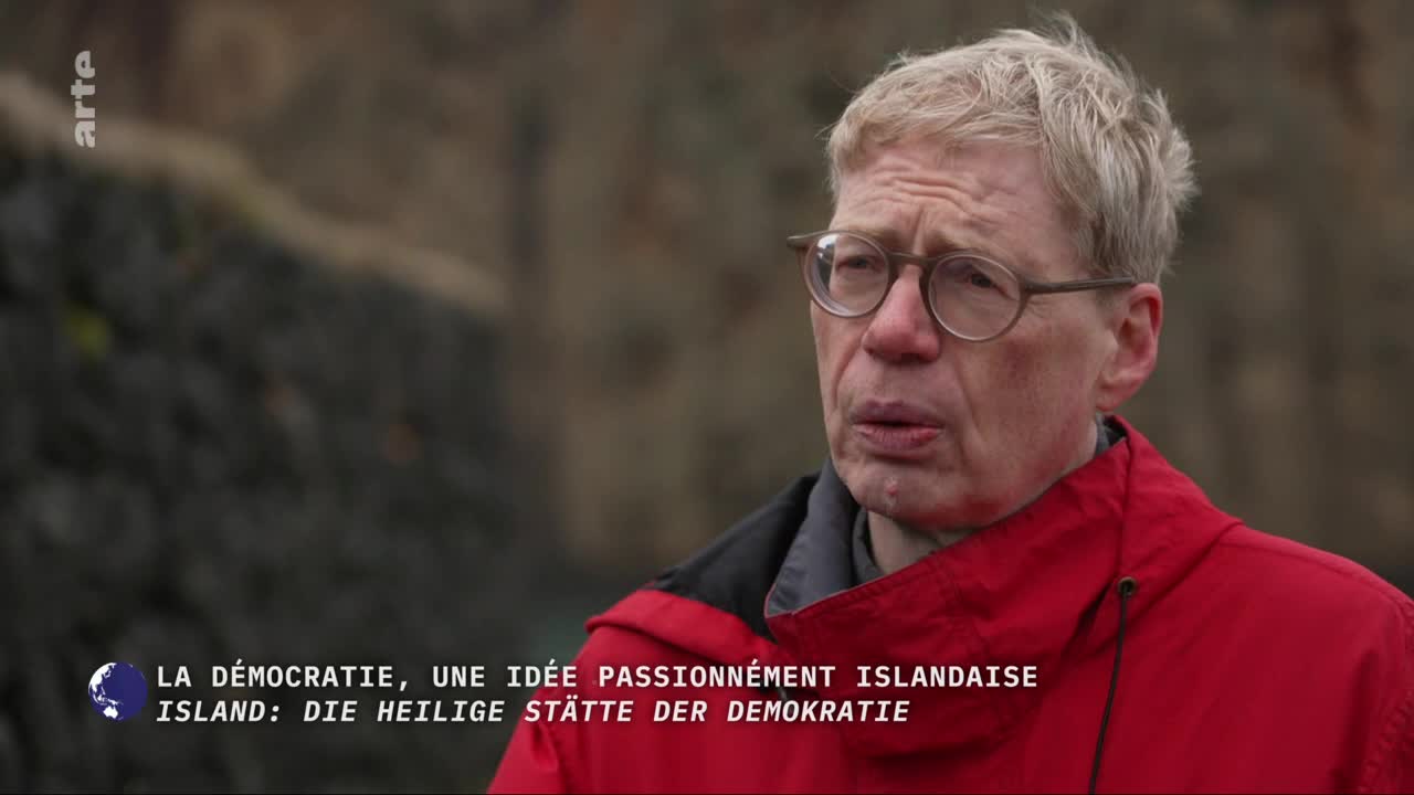 A man with glasses and fair hair speaks, his red jacket bright against the muted, rocky background. The arte logo appears in the upper left corner, and French and German text is displayed at the bottom. A man with glasses and fair hair speaks, his red jacket bright against the muted, rocky background. The arte logo appears in the upper left corner, and French and German text is displayed at the bottom.