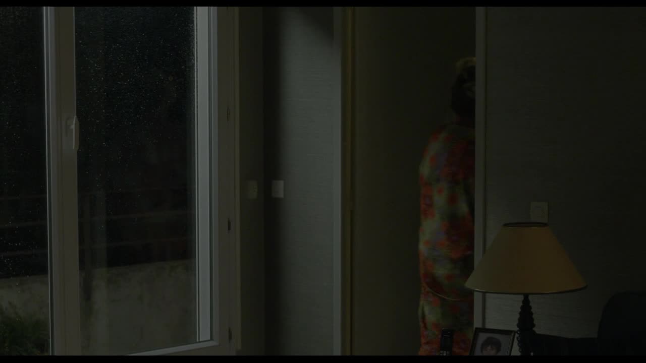 A person in a brightly patterned robe peeks from behind a doorway. Outside, rain streaks down the glass of a large window. A person in a brightly patterned robe peeks from behind a doorway. Outside, rain streaks down the glass of a large window.