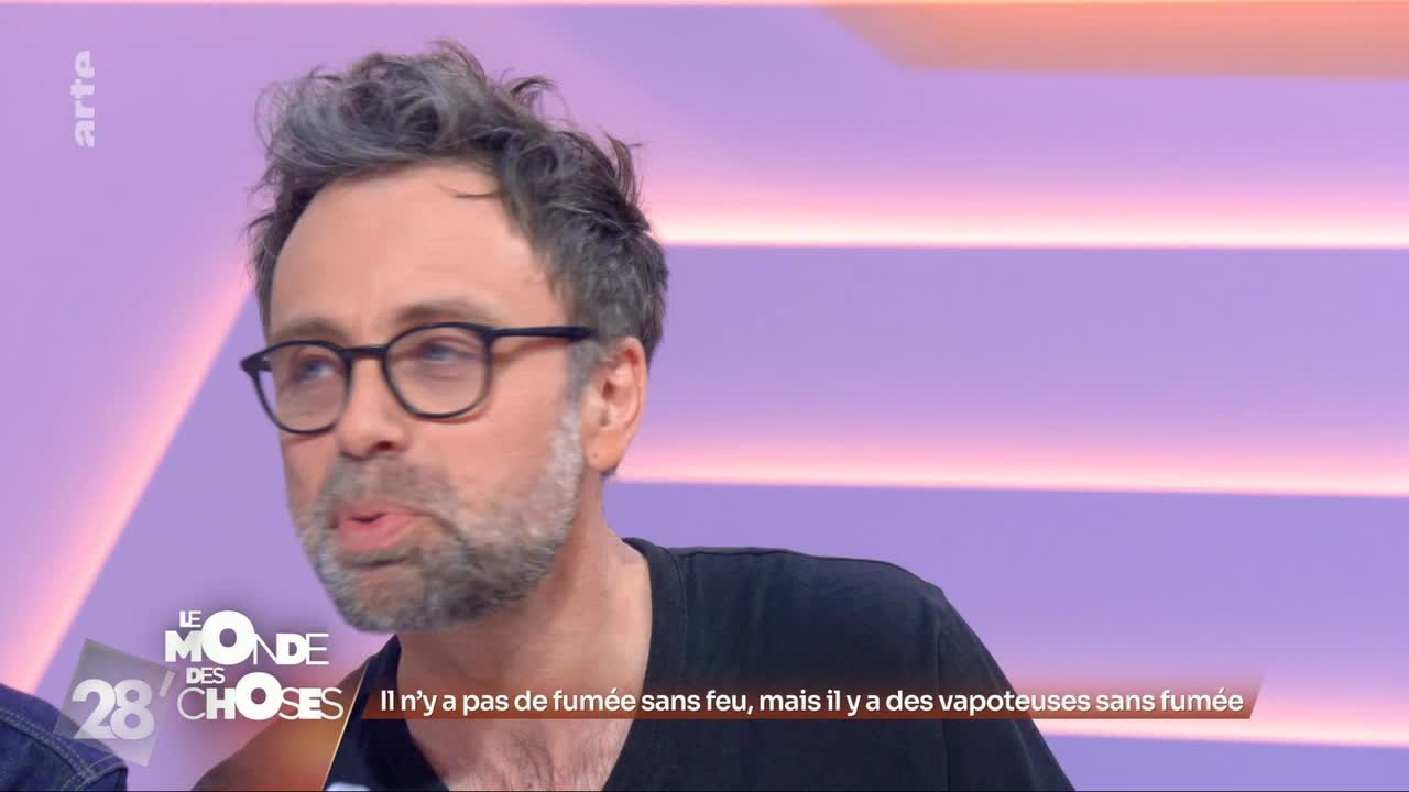 A man with glasses and tousled hair speaks, his mouth slightly open. The backdrop behind him is a soft gradient of purple and pink, with horizontal light bands. A man with glasses and tousled hair speaks, his mouth slightly open. The backdrop behind him is a soft gradient of purple and pink, with horizontal light bands.