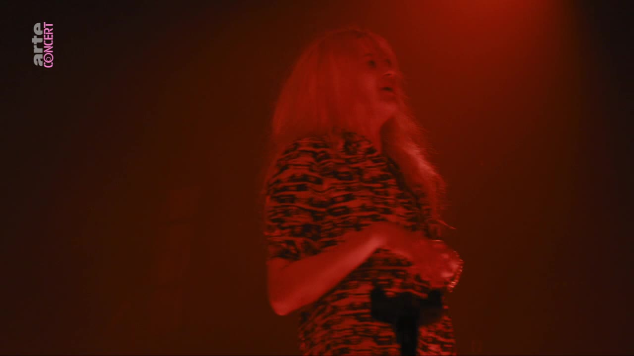 A woman with long blonde hair sings into a microphone, her patterned shirt illuminated by a deep red stage light. The arte concert logo appears in the upper left corner. A woman with long blonde hair sings into a microphone, her patterned shirt illuminated by a deep red stage light. The arte concert logo appears in the upper left corner.