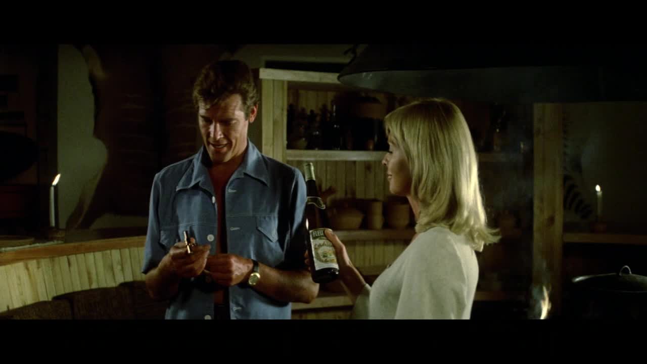 A man in a blue shirt fiddles with a lighter while a woman offers him a bottle. The warm light from a nearby candle casts long shadows. A man in a blue shirt fiddles with a lighter while a woman offers him a bottle. The warm light from a nearby candle casts long shadows.