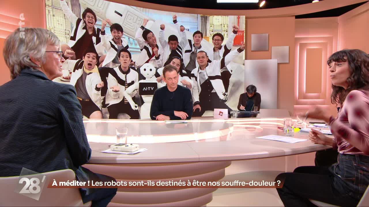 The screen behind the panel shows a group of young men in matching jackets, some cheering. A small, white robot sits among them. On the table in front of me, a glass of water and a small white device rest on a white mat. The screen behind the panel shows a group of young men in matching jackets, some cheering. A small, white robot sits among them. On the table in front of me, a glass of water and a small white device rest on a white mat.