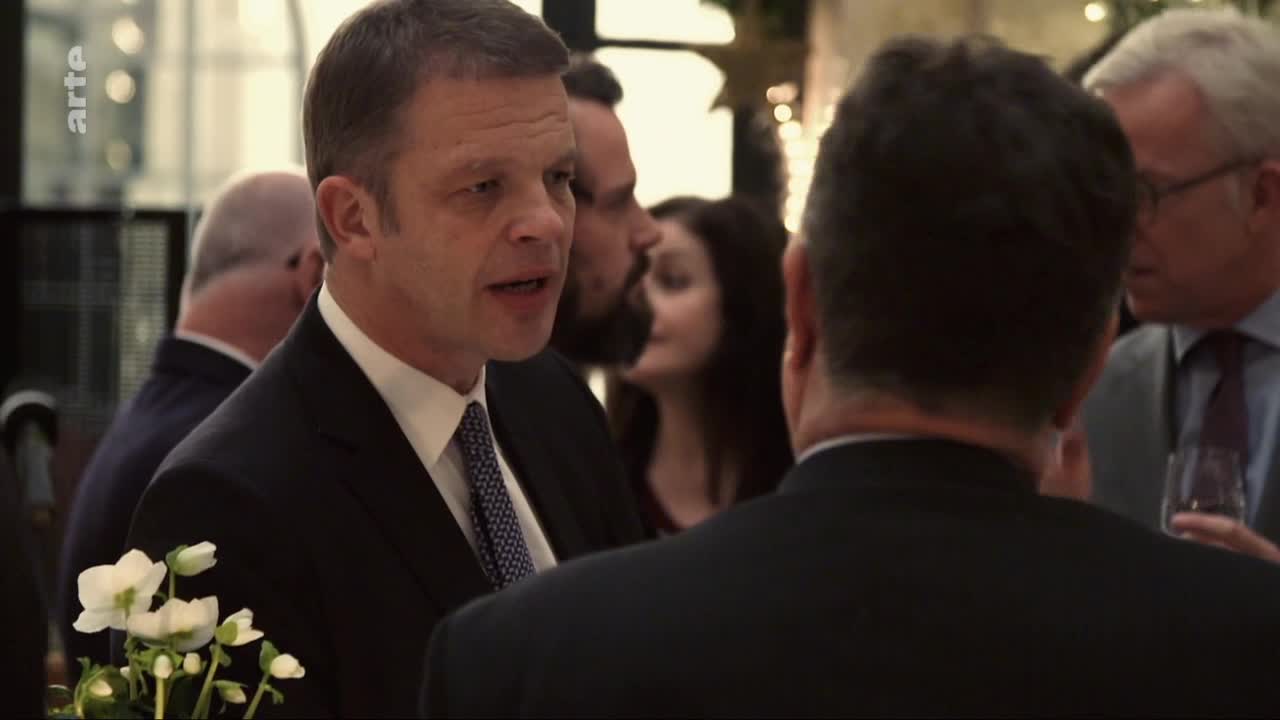 A man in a dark suit and patterned tie speaks animatedly. Behind him, a woman with dark hair smiles faintly. A man in a dark suit and patterned tie speaks animatedly. Behind him, a woman with dark hair smiles faintly.