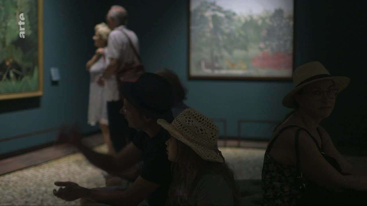 A few people are gathered in a dimly lit gallery, their attention drawn to the art. A woman in a straw hat sits to the right, her gaze directed towards something out of frame. A few people are gathered in a dimly lit gallery, their attention drawn to the art. A woman in a straw hat sits to the right, her gaze directed towards something out of frame.